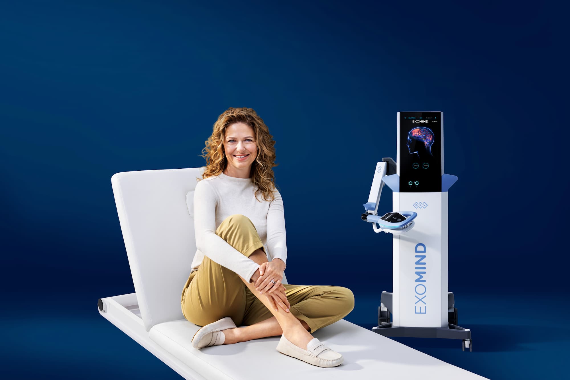 A woman sitting on a treatment bed next to an Exomind device, a treatment which can support a patient's efforts with Body Contouring in Abilene.